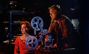 Two people are sitting with a film projector between them. A white woman in red sits on the left whilst a white man in a jacket is poised over the projector, and sits highest among all in the frame. 
