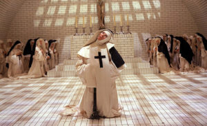 A nun in white with a large black cross emblazoned on the front tilts her head, apparently in a frenzy. 