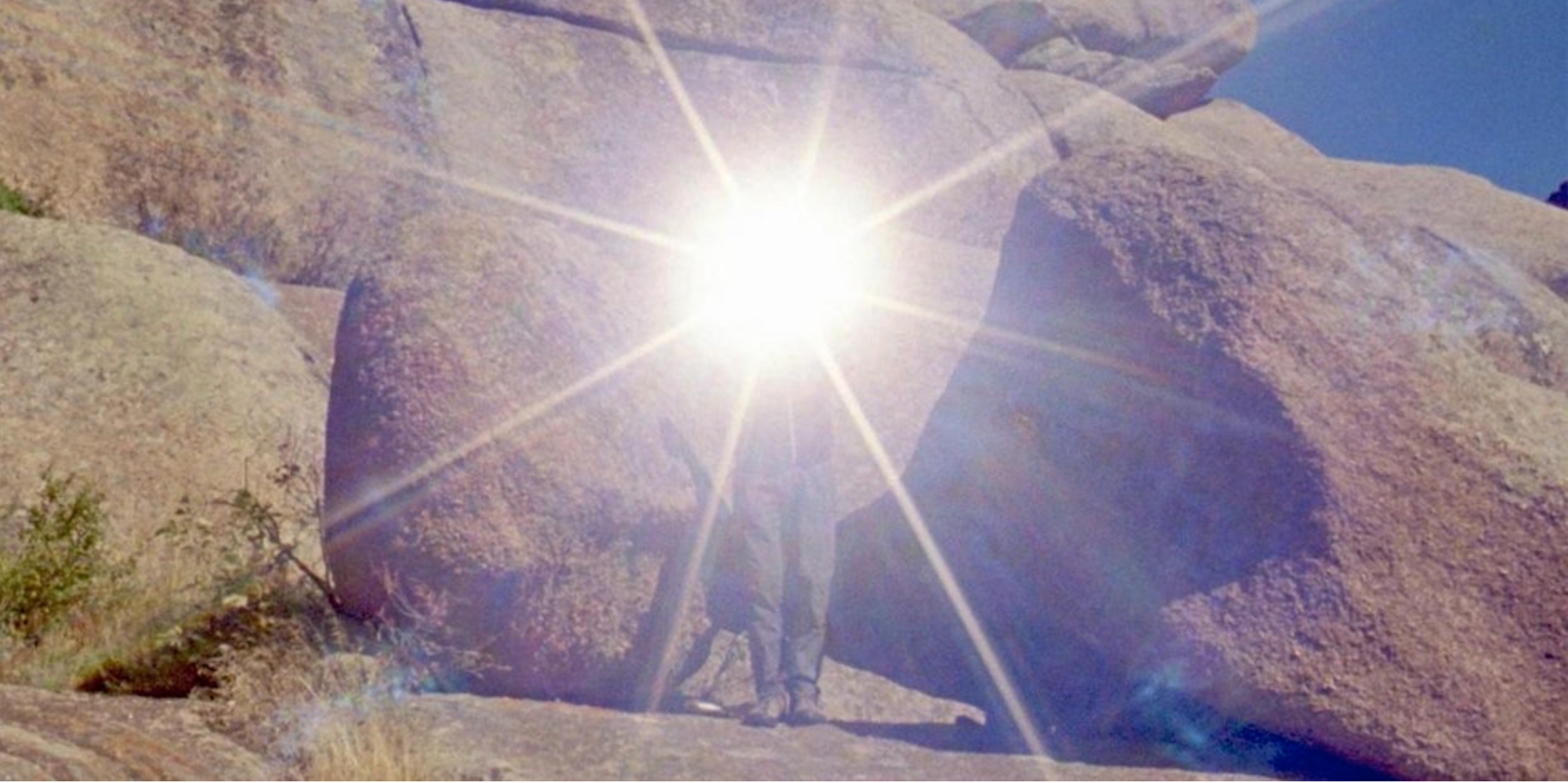 The image shows a﻿ person wearing grey trousers standing with their feet together in front of some very large sandstone rocks. There is a very bright light shining from in front of their face which covers the top half of their body and shines outwards in a star shape.